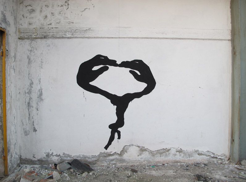 Black painted mural on a rough plastered wall showing two curved arms and hands meeting at the fingertips to form a loop, with a small falling figure hanging from the bottom, rubble scattered on the floor.
