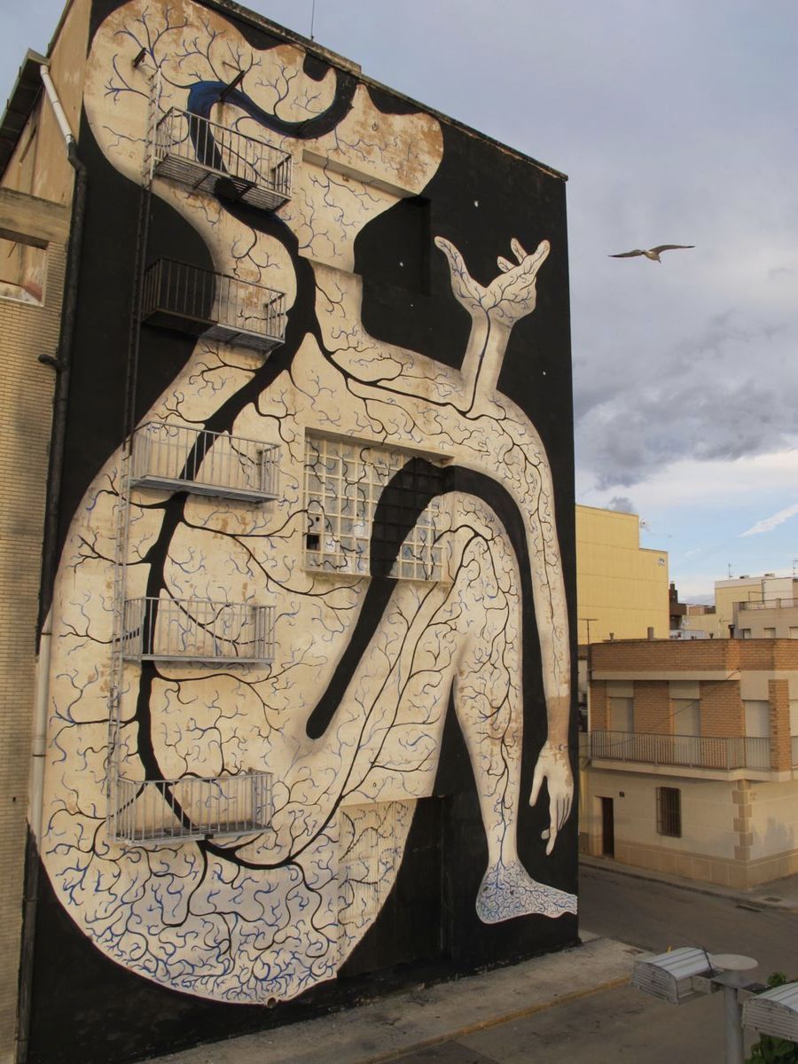 Tall black silhouette figure with a tree-root textured body and a small blue bird held in its curling tail-like limb, painted on the side of a building beside a fire escape, a gull flying past under a cloudy sky.