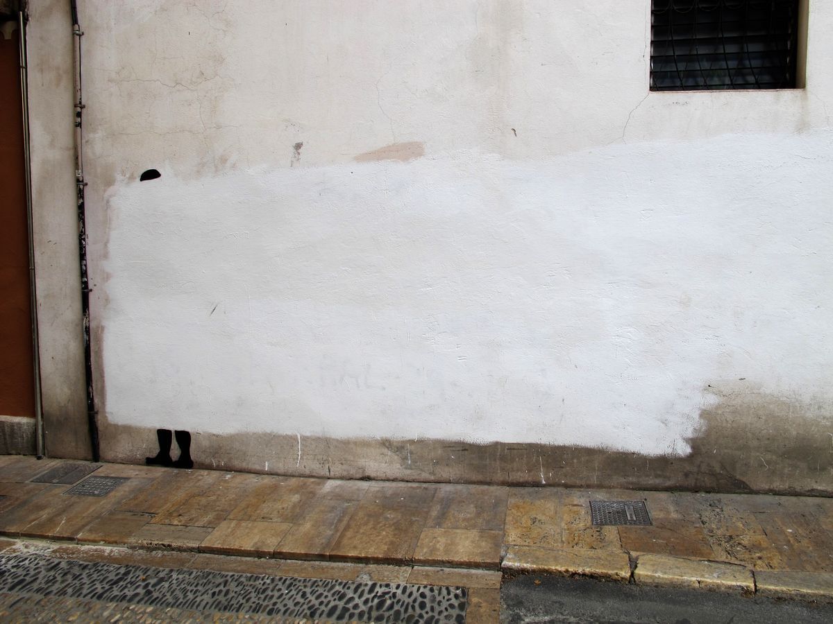 Minimalist mural on a white-painted wall showing only a pair of small black boots and the top of a head peeking up from the bottom edge, as if a figure is hiding behind the wall, a stone sidewalk in front.