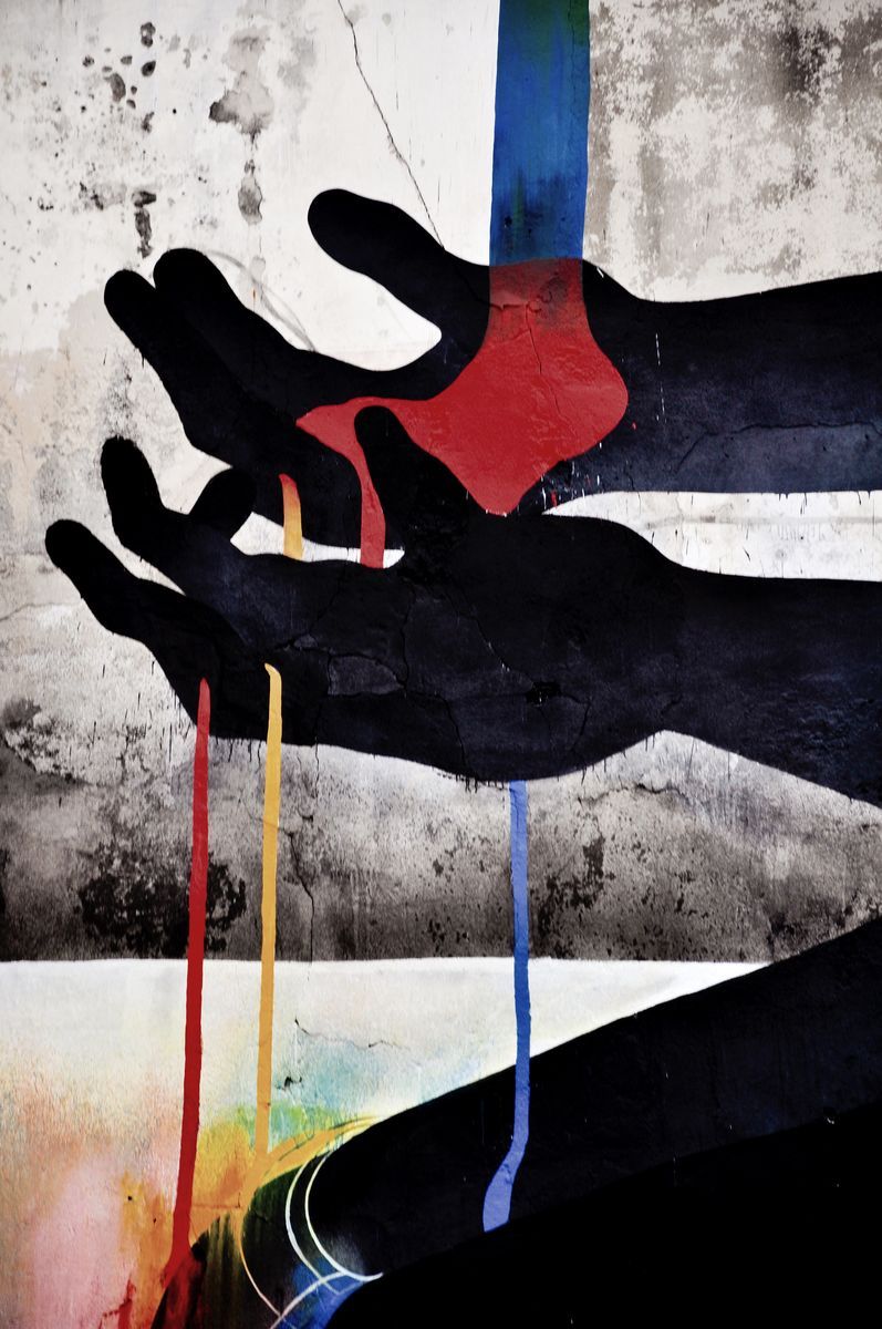 Large black silhouette of a crouching figure on a building facade, head bowed, pouring a rainbow-colored stream from its cupped hands down toward a row of advertising billboards, a car driving past on the street below.