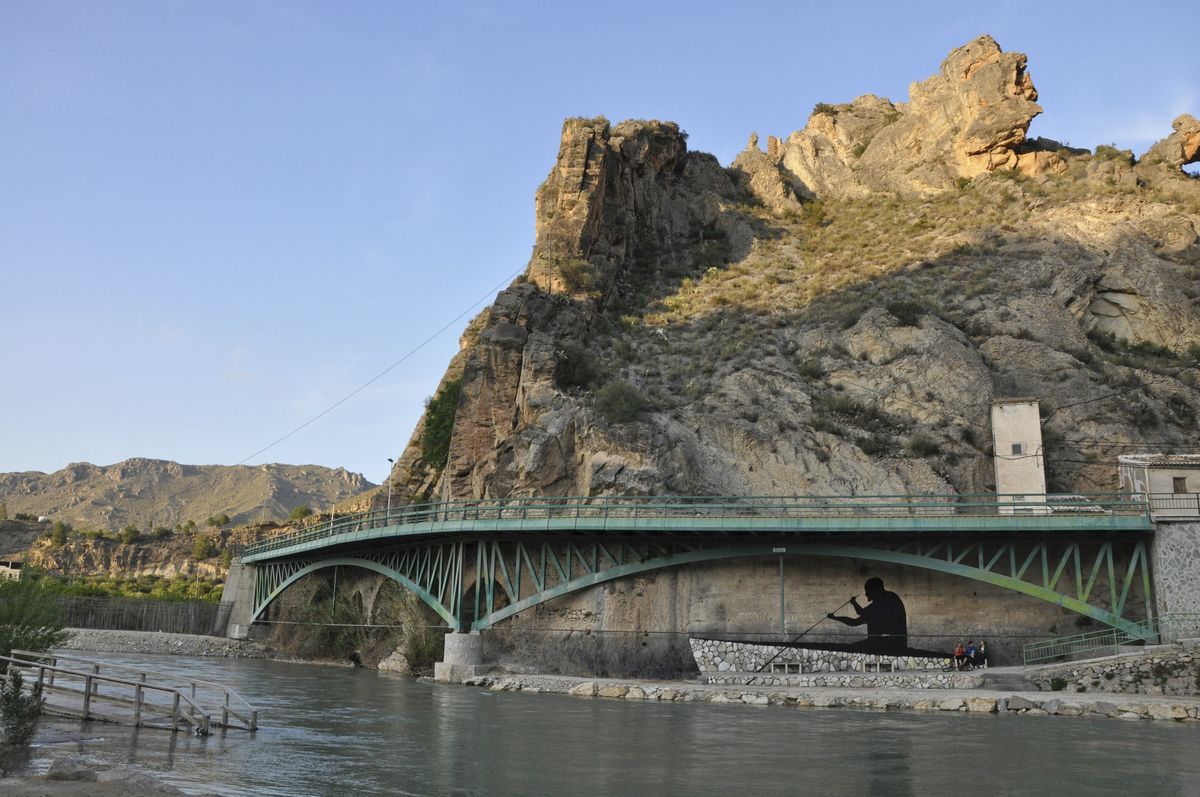 Wide view of a green steel arch bridge crossing a river beneath a rocky cliff, a black silhouette of a man rowing a boat painted on the stone wall under the bridge.