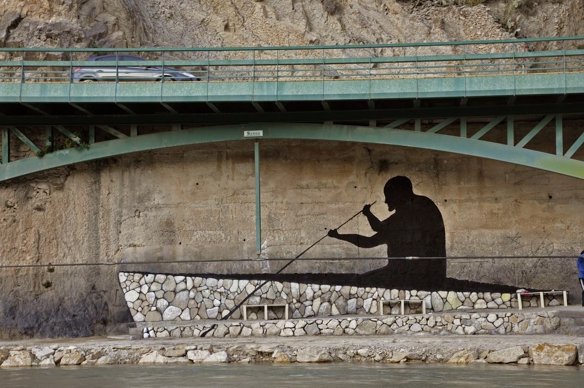 Closer view under the bridge of the black silhouette mural showing a seated man rowing with a long oar in a boat shape built from stacked stones, a car crossing the bridge above.