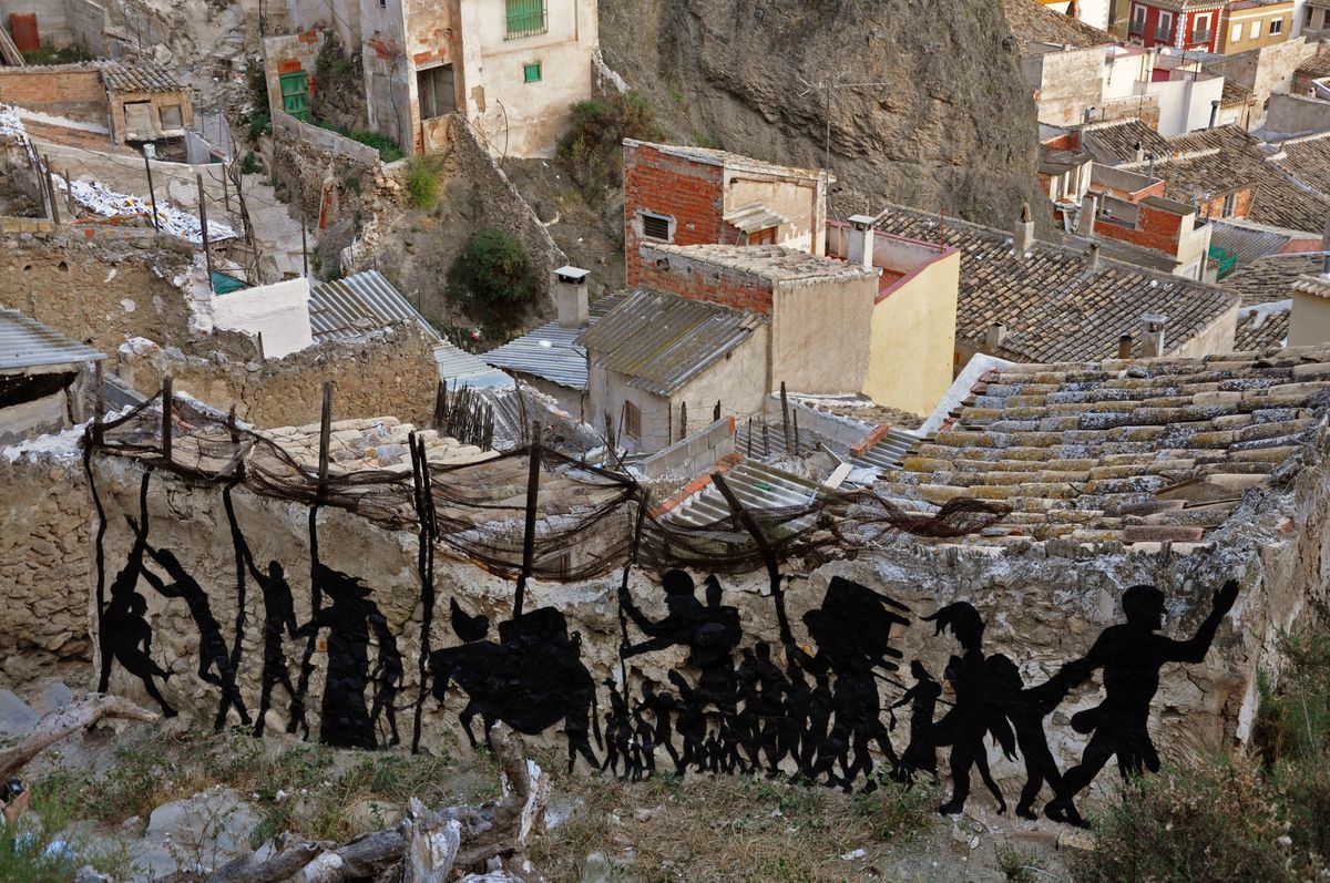 Black silhouette mural of a long procession of people and a donkey with baskets, walking single file across a ruined rooftop, an old fishing net strung above them, a hillside village visible behind.