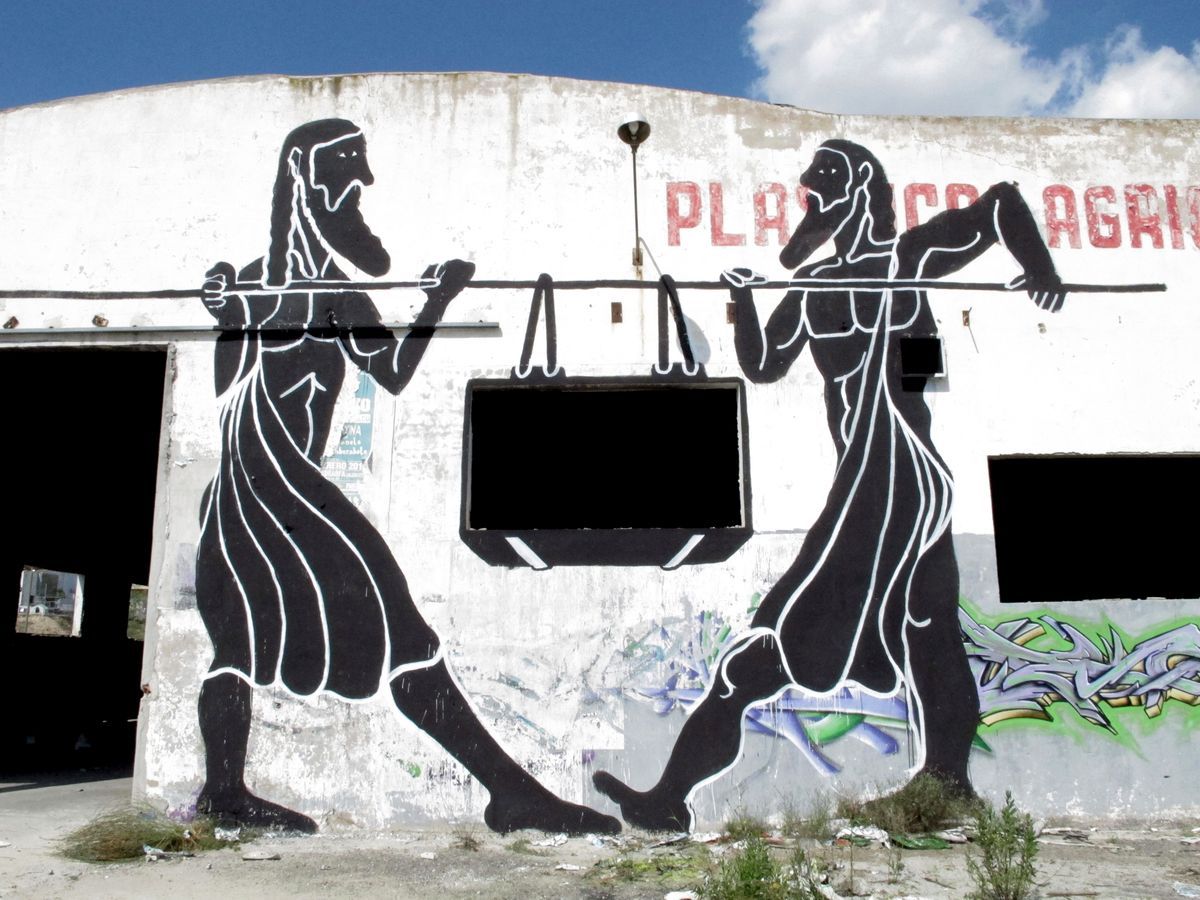 Two black silhouette figures in long robes carrying a pole across their shoulders with a box hanging from it, painted on the facade of an abandoned warehouse, graffiti tags on the wall beside them.