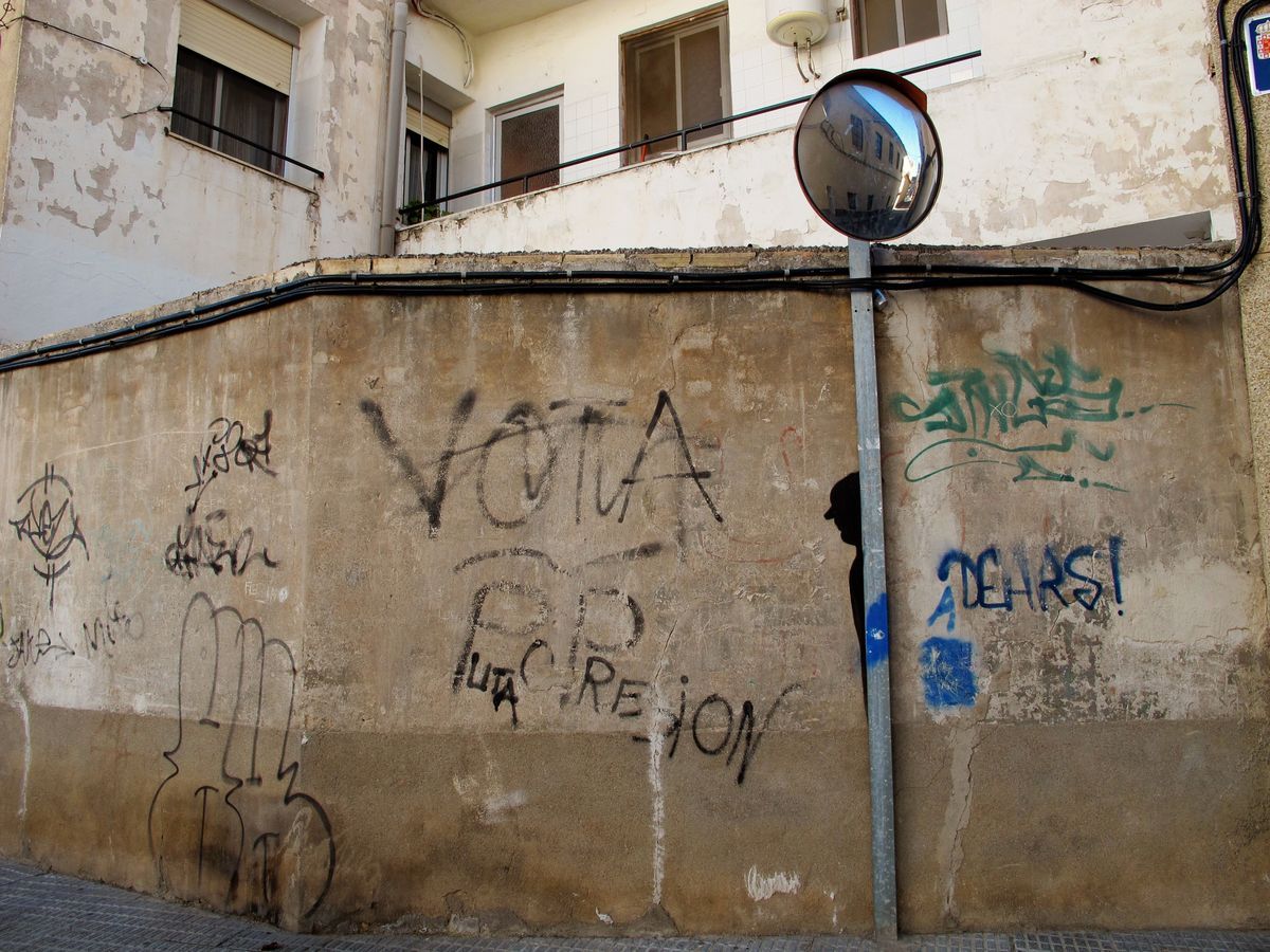 Weathered concrete wall covered in scattered graffiti tags and political slogans, a black silhouette figure blending into the wall beside a street pole with a convex traffic mirror.