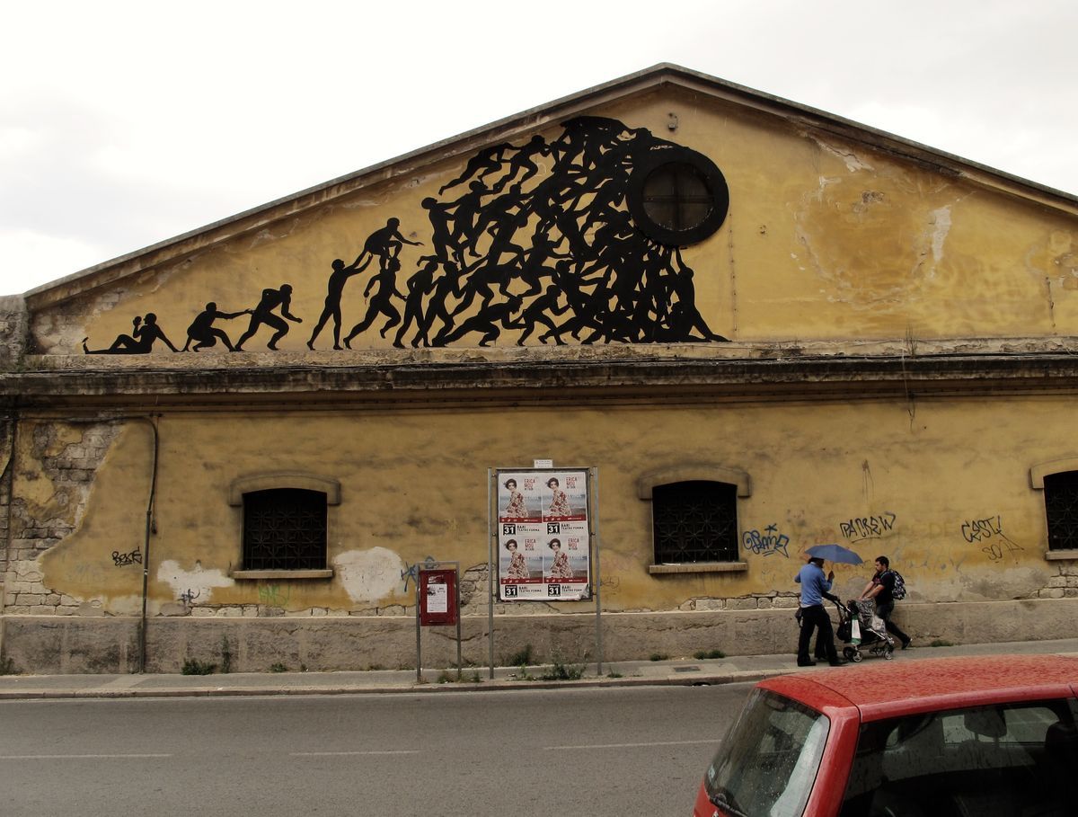 Wide view of a yellow gable facade with a black silhouette mural of a chain of climbing figures piling toward a round window, two pedestrians with a stroller and a red car passing on the street below.