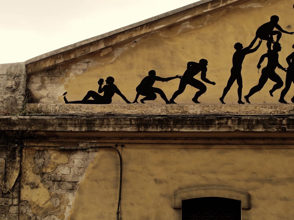 Close-up of the same yellow facade mural showing the chain of black silhouette figures pulling and climbing over one another up the sloped roofline.