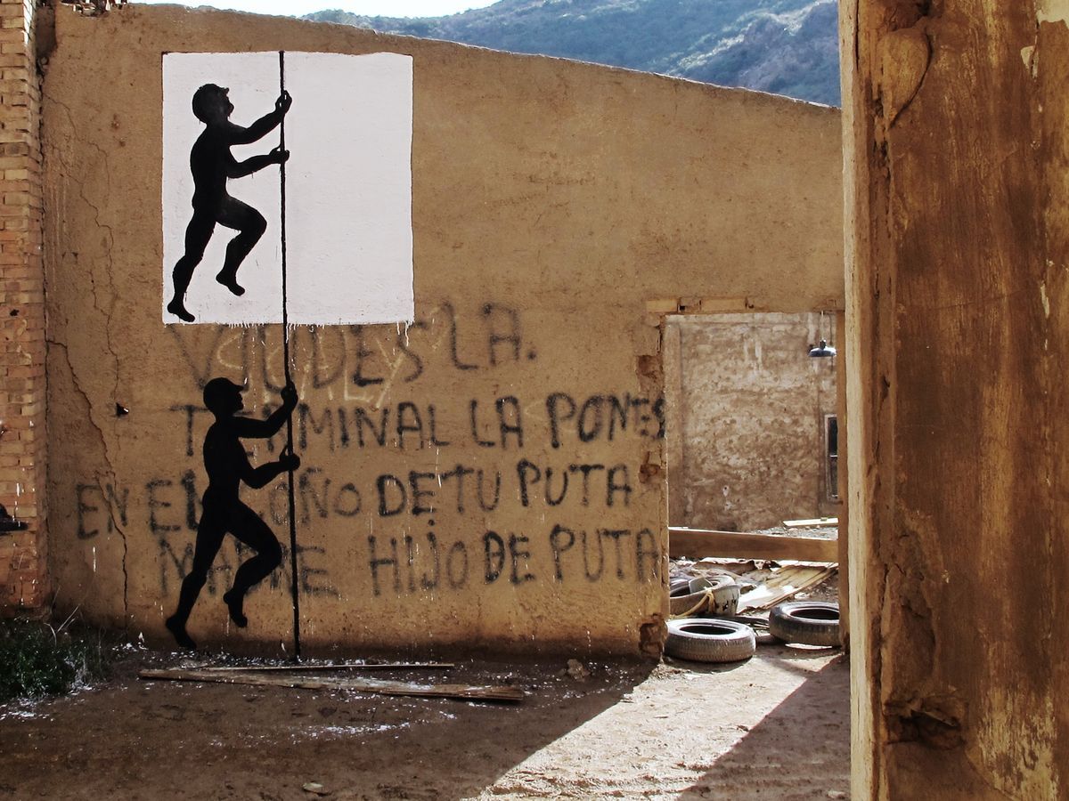 Two identical black silhouette figures climbing a vertical rope on a ruined building wall, one framed by a painted white rectangle, spray-painted graffiti text covering the rest of the wall.