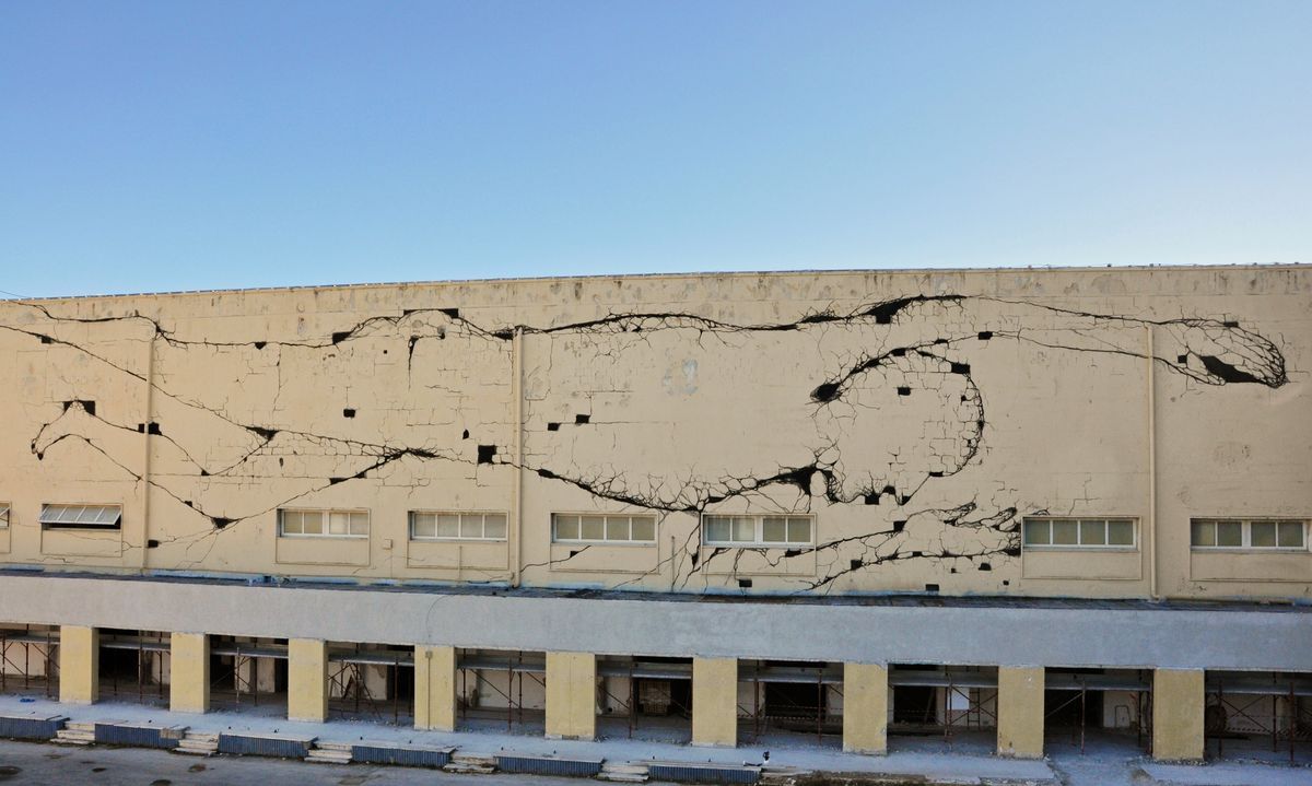 Long building facade painted with black crack-like branching lines resembling roots or veins spreading across the cream-colored wall above a row of windows and an arcade.