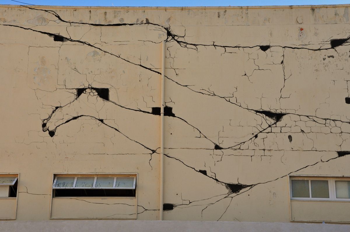 Another section of the crack mural showing black branching lines crossing diagonally across the cream wall above three windows.