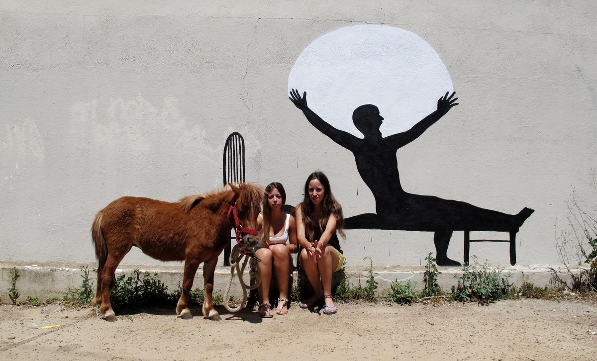 Black silhouette mural of a man on a bench stretching his arms wide before a large white sun or moon circle, painted on a wall, two women and a small pony sitting in front of it.