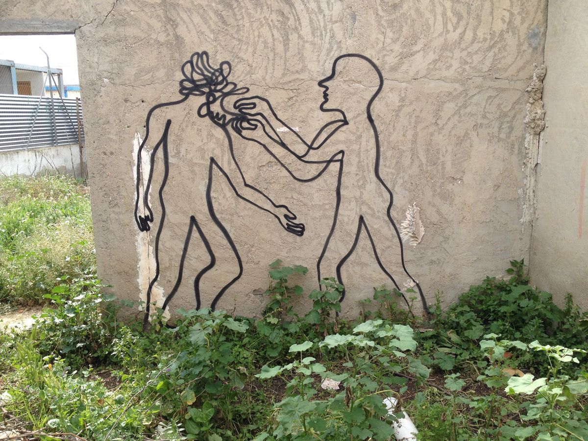 Two black outline figures spray-painted on a rough plastered wall, one combing the other's scribbled hair, surrounded by overgrown weeds and a corrugated metal fence.