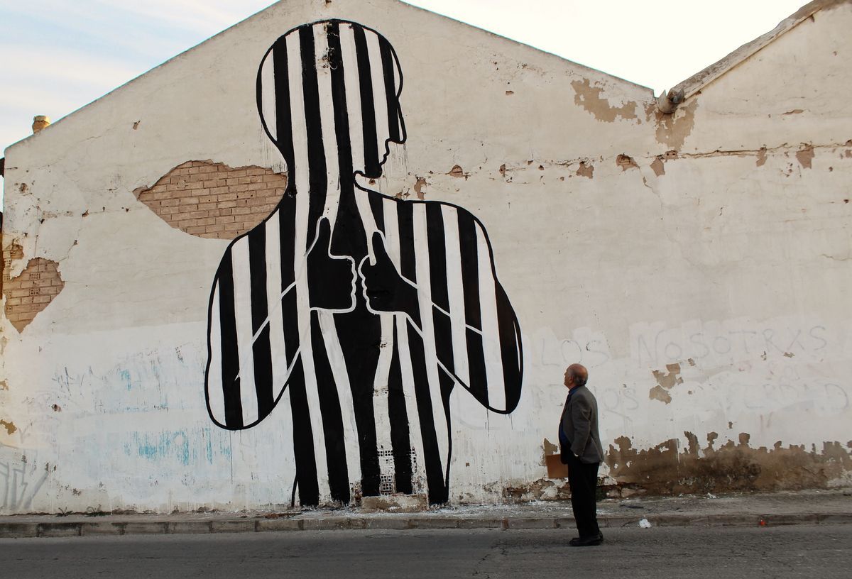 Black and white striped silhouette of a standing figure with two thumbs-up hands crossed over its chest, painted on a weathered gable wall, a man standing below looking up at it.