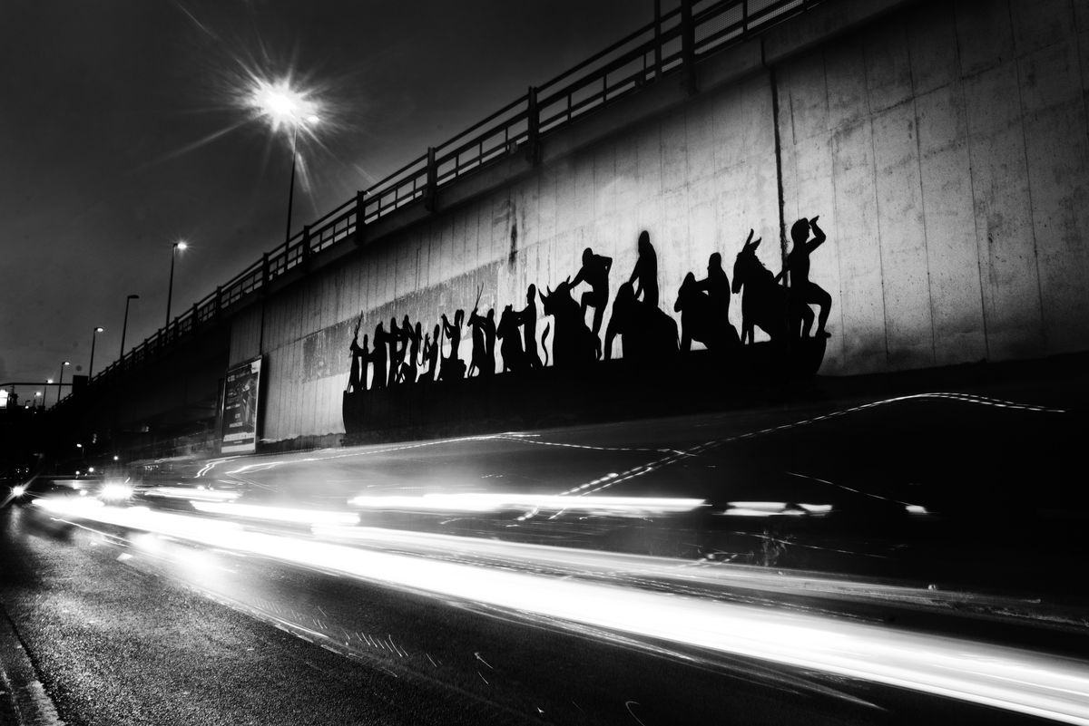 Black silhouette mural of a boat crowded with people and animals holding hands in a row, its reflection painted below on a concrete wall beneath an overpass.