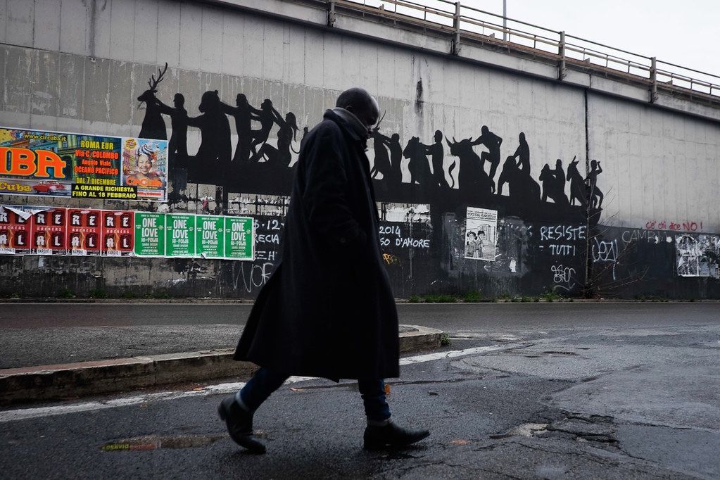 Black silhouette mural of a chain of figures and a deer holding hands along a raised concrete ledge, a worker on a hoist painting the piece while two pedestrians walk past below.