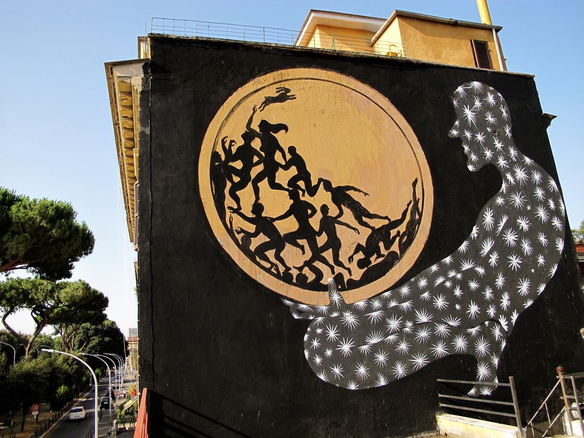Black silhouette mural on a building corner showing a circle of dancing figures with a leaping rabbit, beside a large starburst-patterned humanoid silhouette, trees and a street lined with lampposts below.