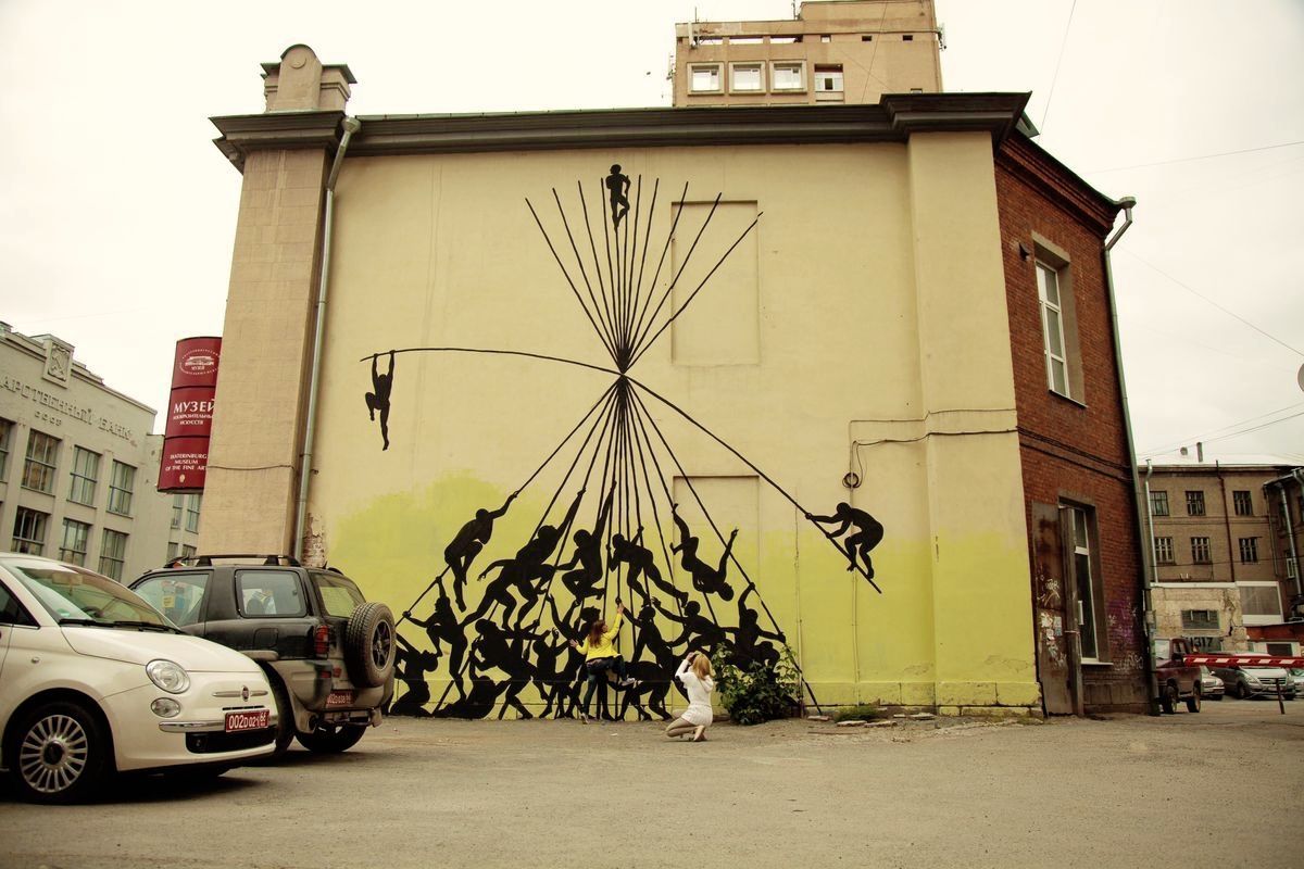 Wide view of a building facade mural showing a triangular arrangement of black silhouette figures climbing a web of ropes toward a peak, two people photographing the mural from the parking lot below.