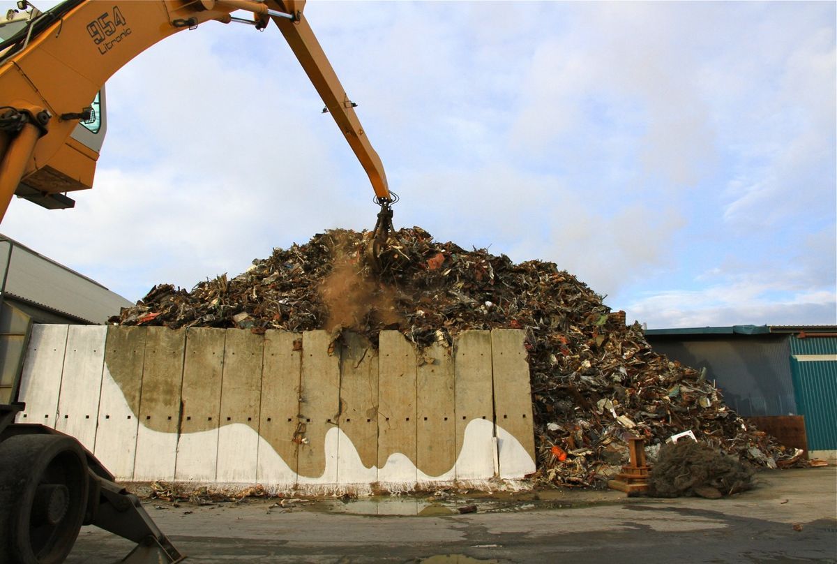 Excavator with a grapple claw dropping scrap metal onto a large pile beside a concrete barrier wall painted with a white wave-shaped mural, an industrial scrapyard setting.