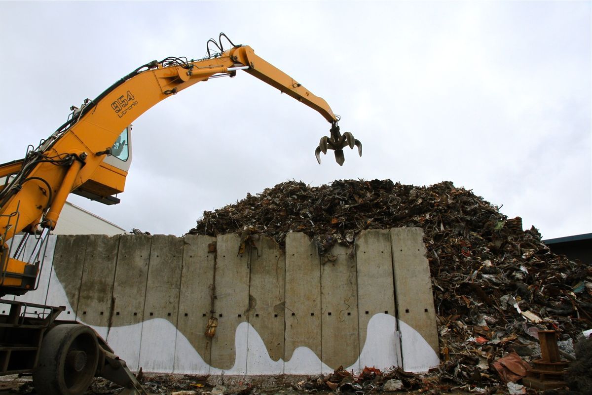 Low-angle view of the same scrapyard, the excavator's claw raised over a mountain of scrap metal above a concrete barrier painted with a white wave pattern.
