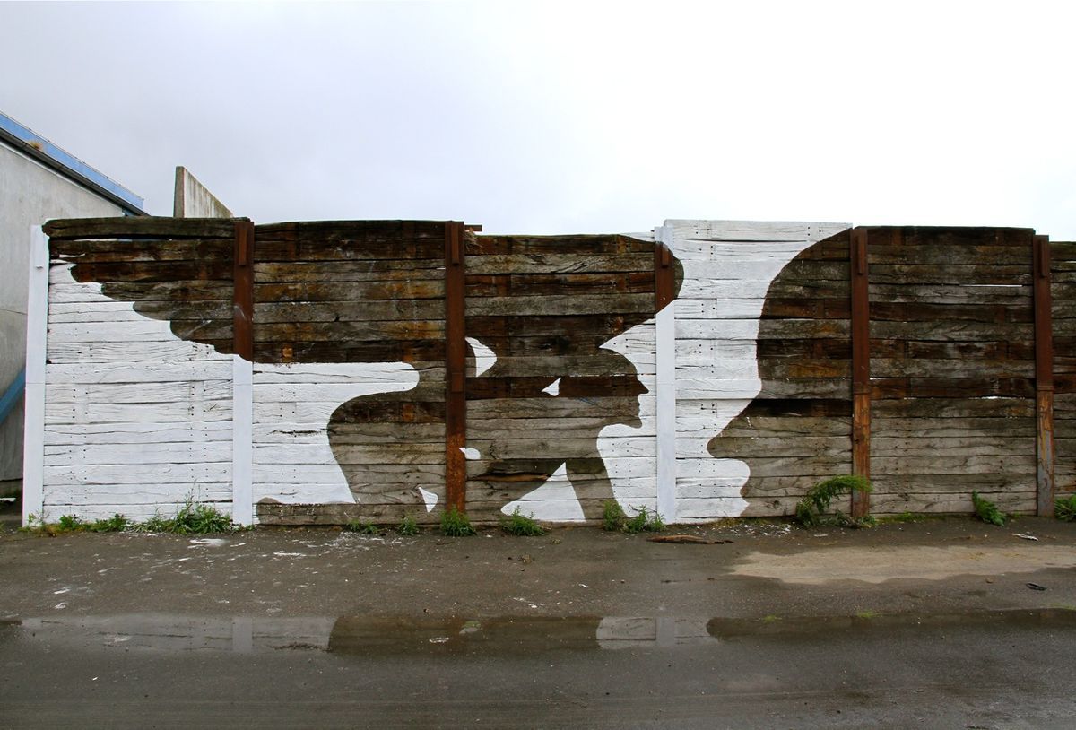 Two black-and-white profile heads facing each other painted across a row of weathered wooden fence panels, one with a wing shape extending from its head, a wet street reflecting the mural below.