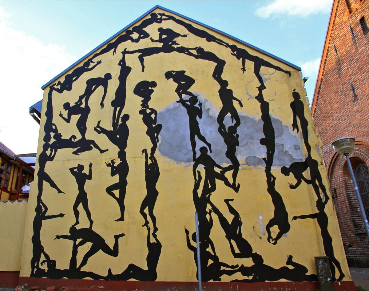 Yellow gable wall covered in a dense black silhouette mural of dozens of interlocking human figures forming a grid-like pattern, a brick church tower beside it.