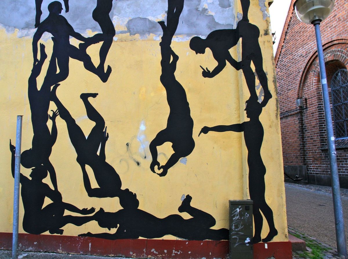 Close-up section of the same yellow wall mural showing black silhouette figures in acrobatic poses, hanging, pointing, and balancing on one another, a lamppost and utility box in the foreground.