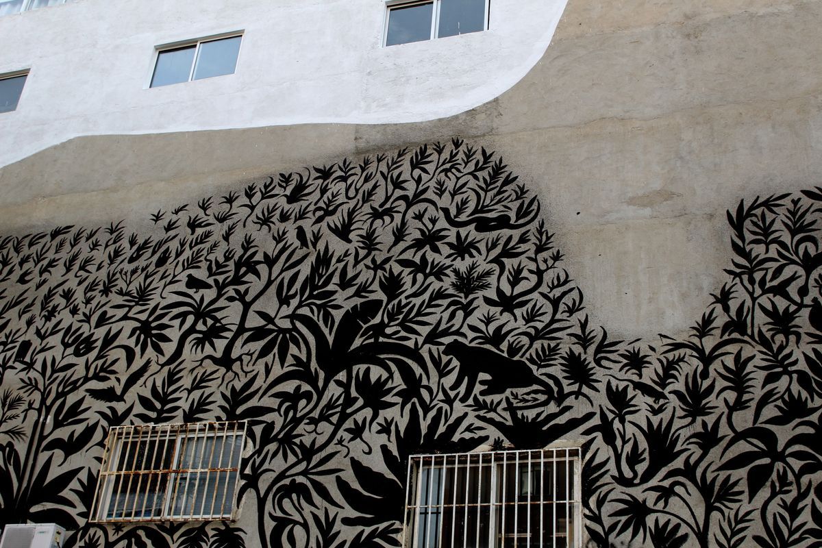 Dense black silhouette mural of leaves and foliage covering a building wall, with hidden animal and bird shapes woven into the pattern, barred windows at the base.