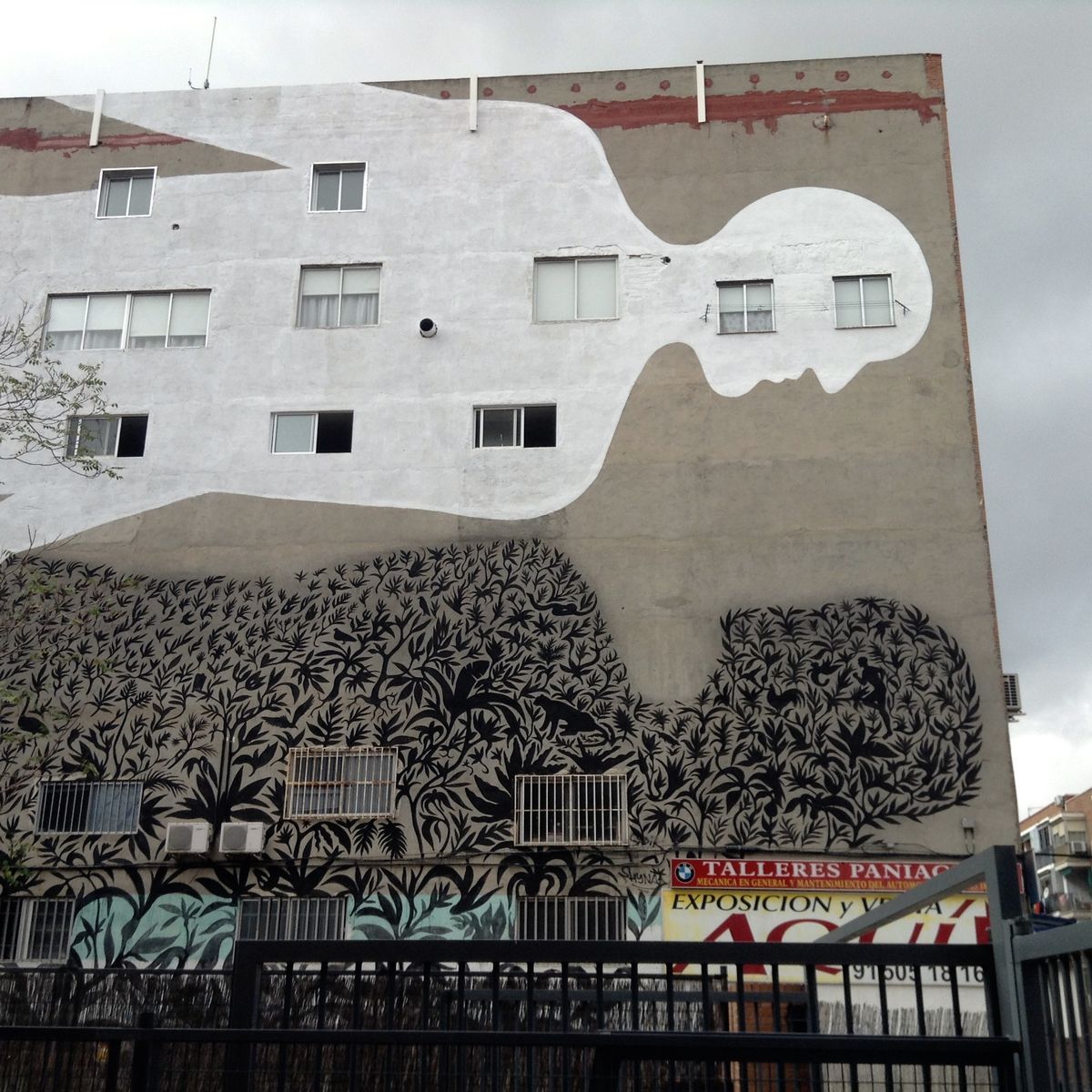 Building corner mural split between a white humanoid silhouette with windows as eyes on the upper floors and dense black foliage on the lower floors, commercial signage visible at street level.