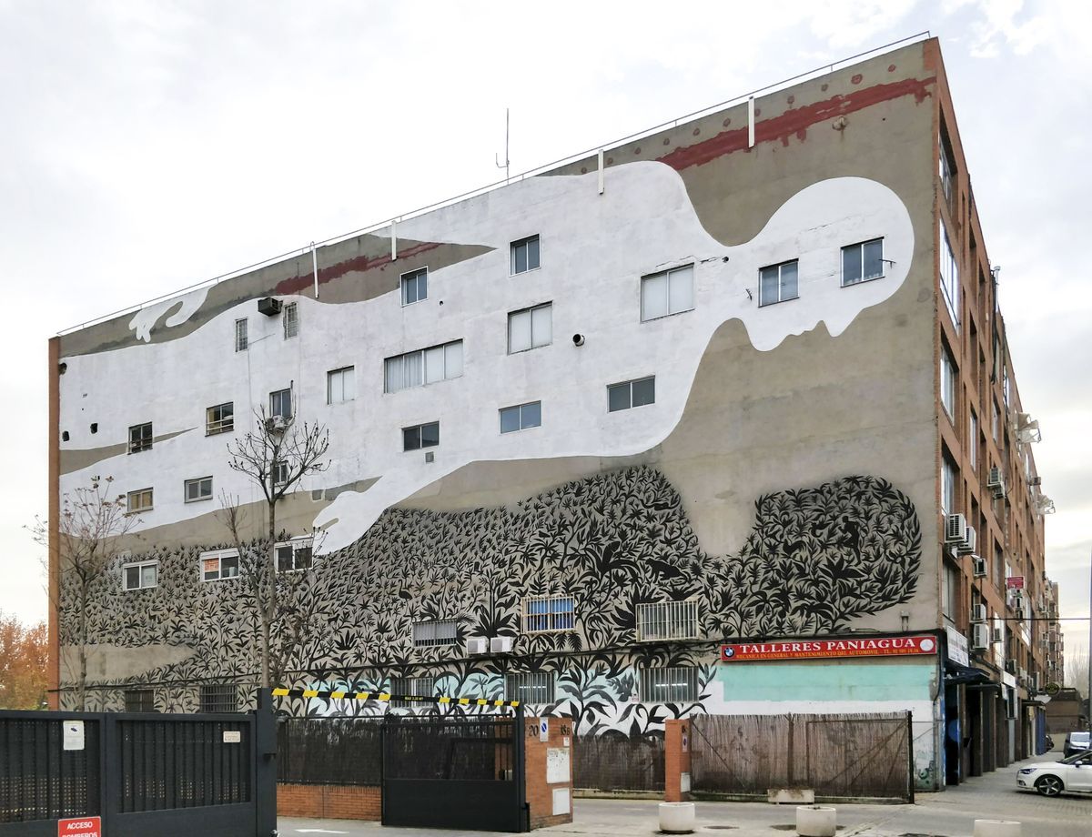 Wide street view of the full building mural showing a white reclining figure spanning the upper stories above dense black foliage on the lower floors, parked cars and shop signs at ground level.