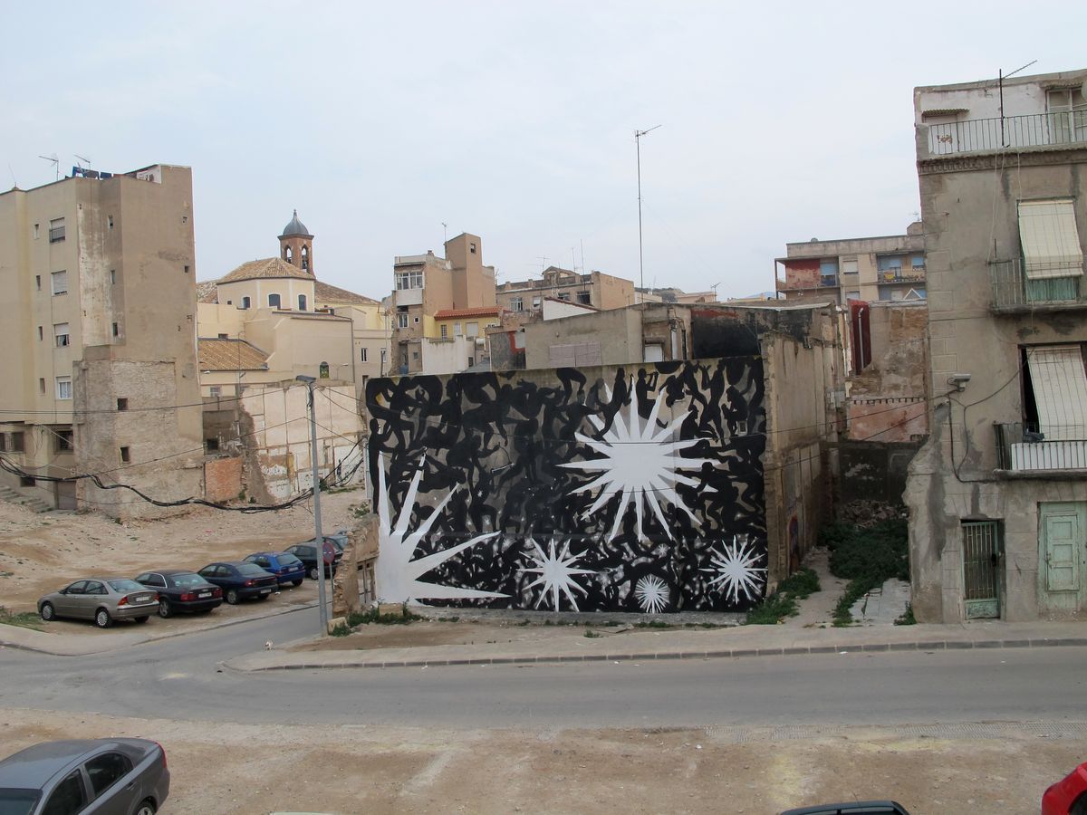 Black silhouette mural of a dancing crowd of figures beneath white multi-pointed starburst shapes, painted on a freestanding wall among ruined buildings, cars parked on the street below.