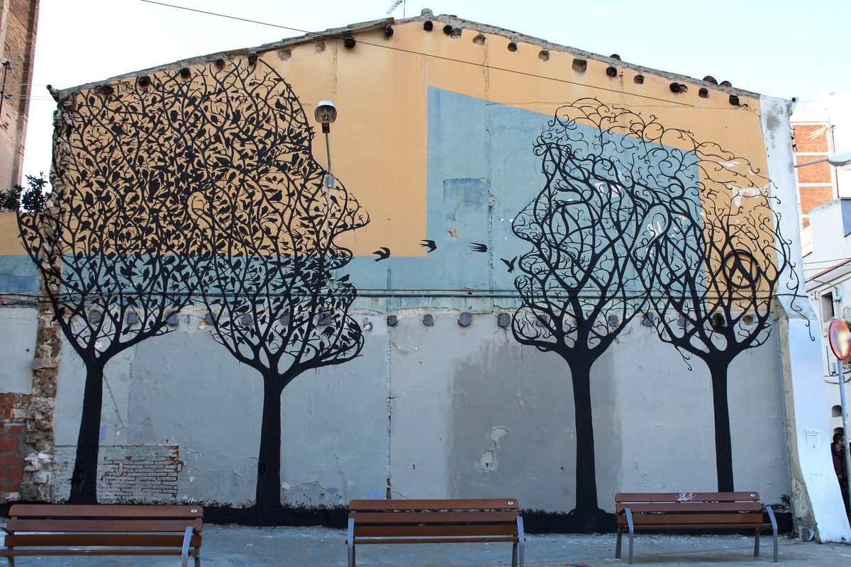 Wide view of a building gable mural showing four black silhouette trees, the leftmost two formed from dense bird-covered branches merging into a human profile, park benches lined up in front.
