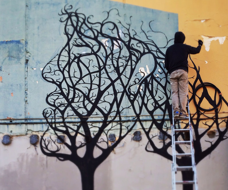 A person on a ladder painting the black branching outline of a tree mural onto a wall, part of the design already filled in below against a pale blue and tan background.