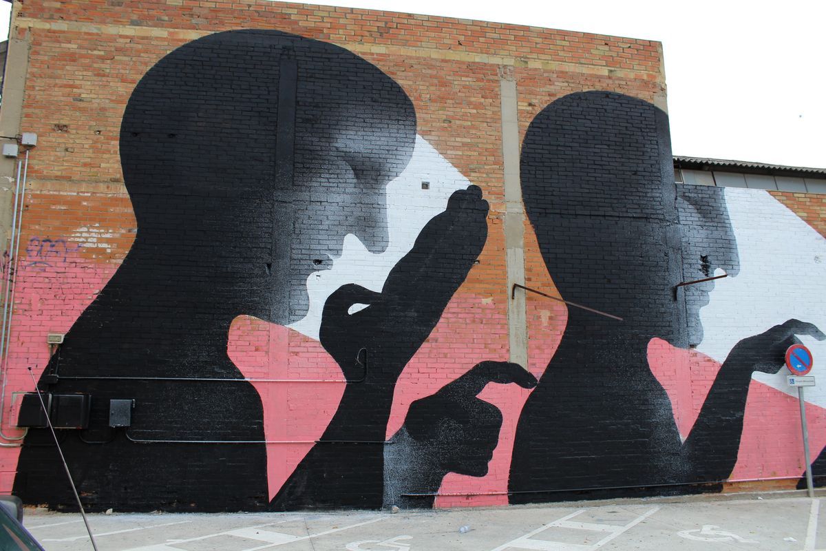 Wide mural on a brick warehouse wall showing two large black silhouette profile heads with raised hands, set against pink and white geometric shapes, a no-parking sign and car mirror visible in the foreground.