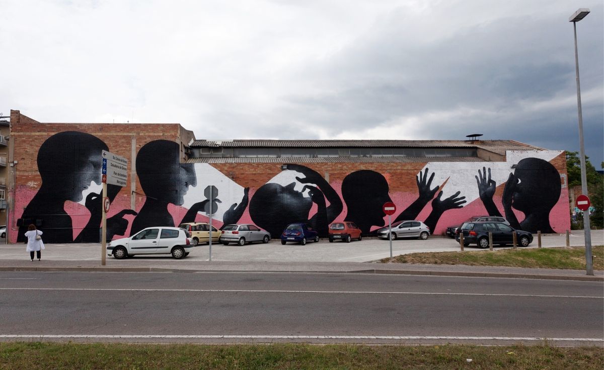 Wide mural along a brick and white-painted warehouse wall showing a row of black silhouette profile heads and raised hands against pink and white geometric shapes, a parking lot with several cars in front and a street sign visible.