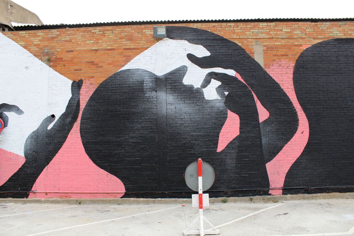 Close-up of the same mural showing two black silhouette heads close together with hands raised near their faces, painted directly on exposed brick, a road sign post in the foreground.