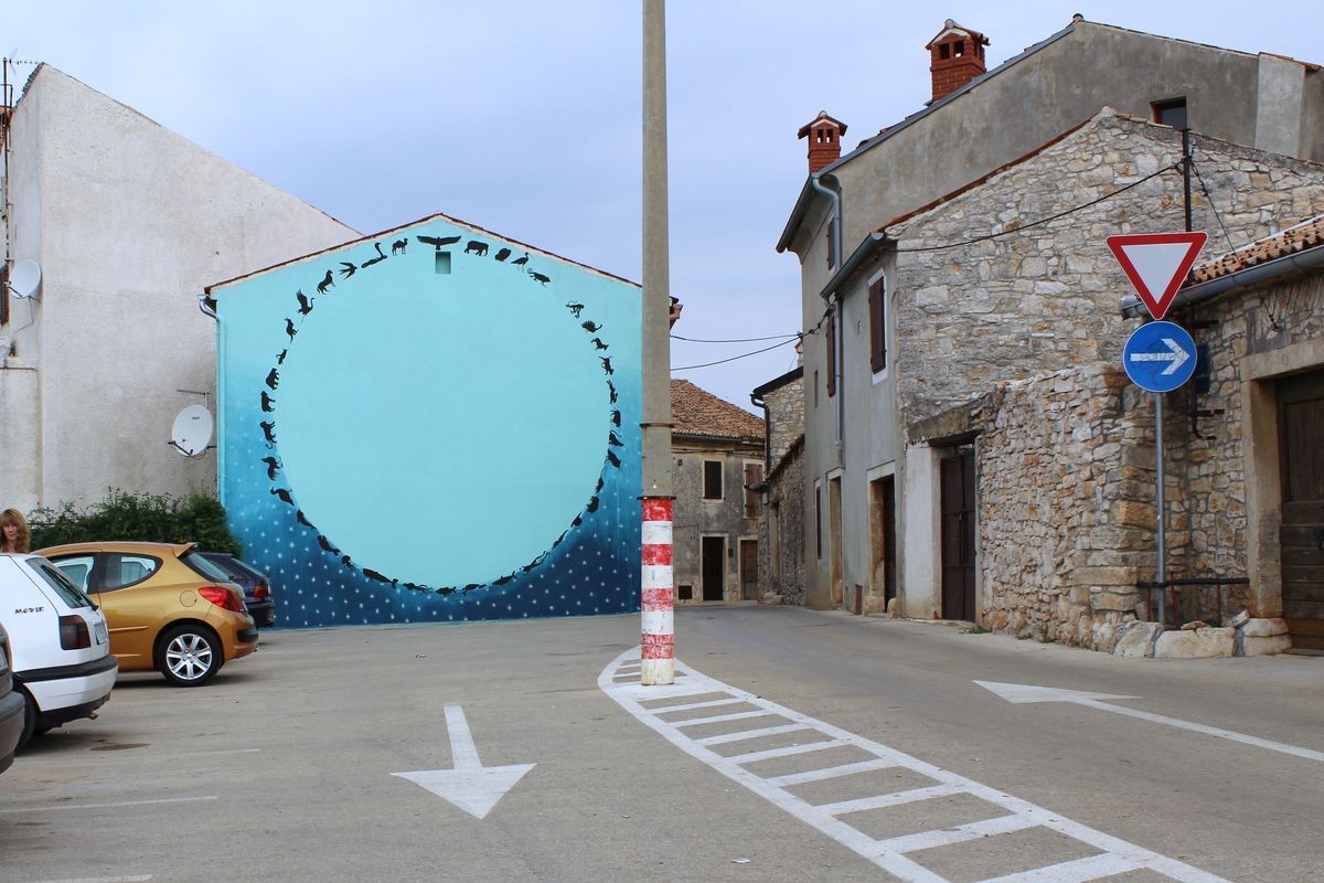 Mural on the gable end of a stone house showing a large circle of small black silhouette animals arranged nose to tail against a blue gradient sky and starry night ground, seen from a street intersection with road signs and parked cars.