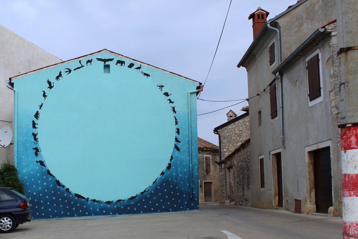 Mural on the gable end of a stone house showing a large circle of small black silhouette animals arranged nose to tail against a blue gradient sky and starry night ground, painted above a narrow street lined with old buildings.