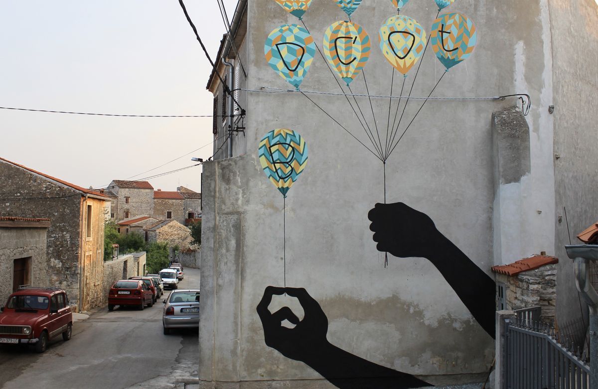 Mural on a building corner showing two black silhouette hands holding strings tied to five patterned hot air balloons, each balloon printed with a single letter, painted above a street lined with parked cars.