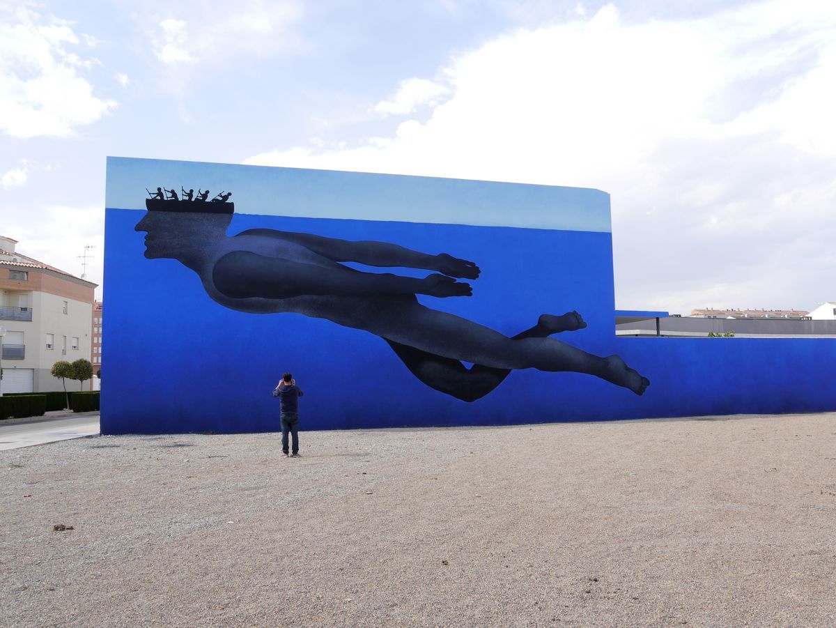 Wide view of a large mural on a blue wall showing the black silhouette of a swimmer gliding underwater, their head formed from a rowing boat crew at the surface, a person photographing the mural from the gravel lot below.