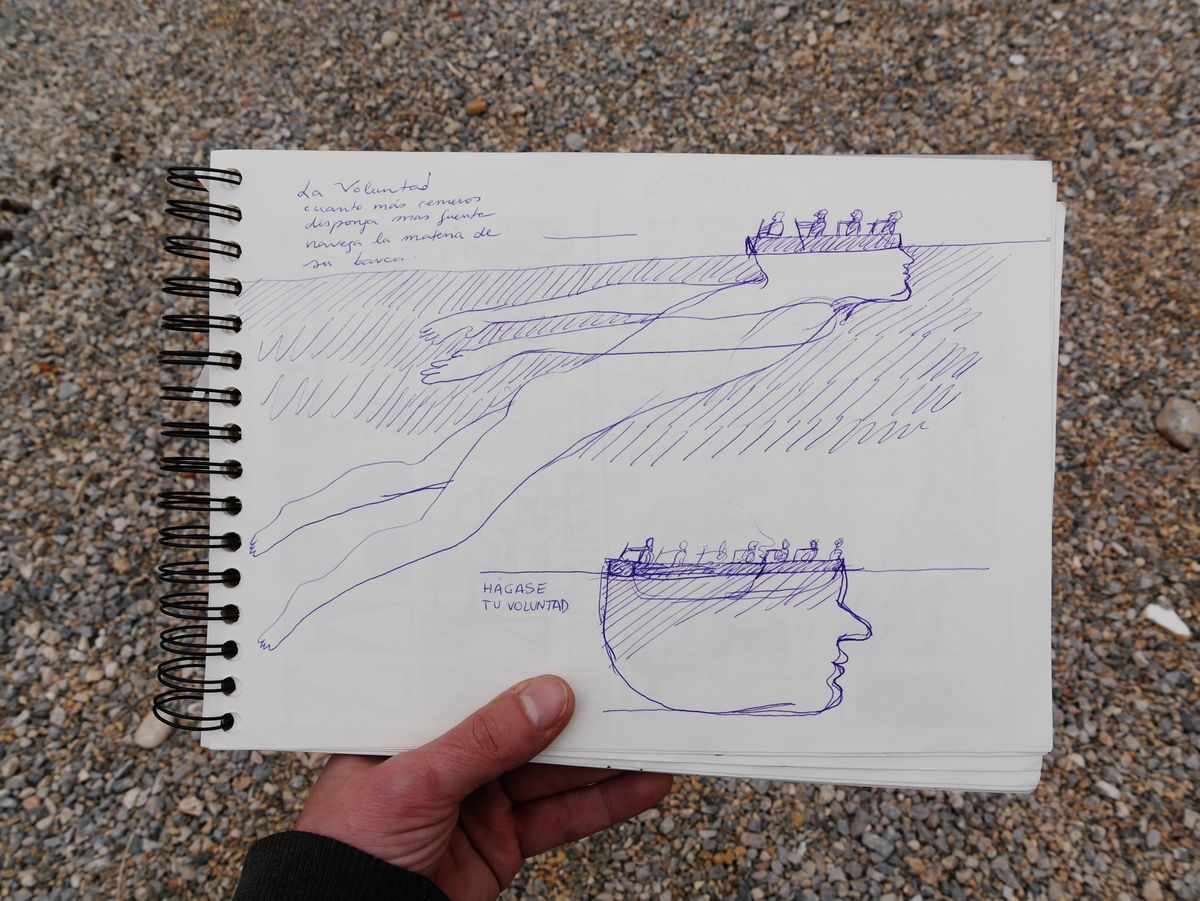 Notebook sketch in blue ballpoint pen showing two versions of the swimmer-with-rowers design, annotated with handwritten Spanish text about willpower, held open in a hand over gravel ground.