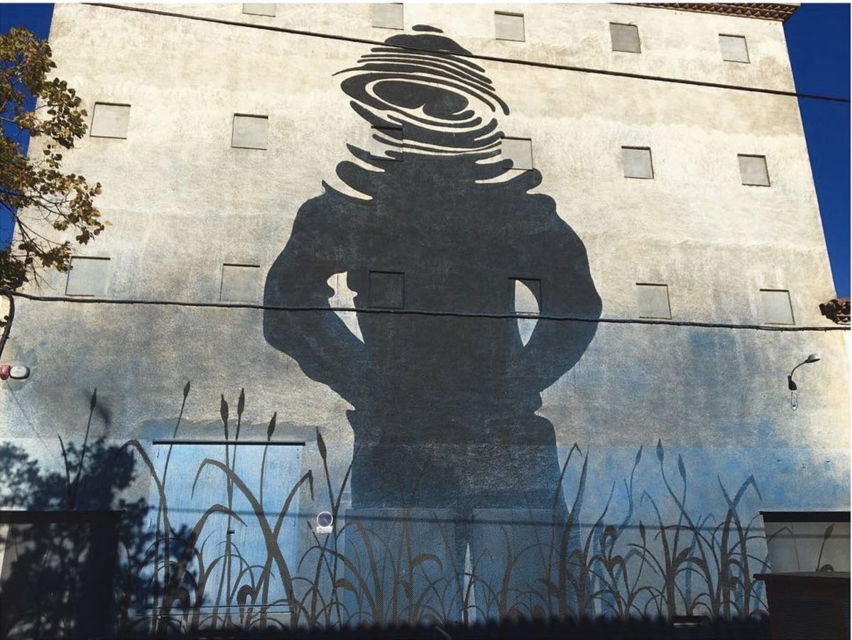 Black silhouette mural of a giant standing figure with hands on hips, its head replaced by concentric rippling water rings, painted on a tall building above rows of painted reeds.