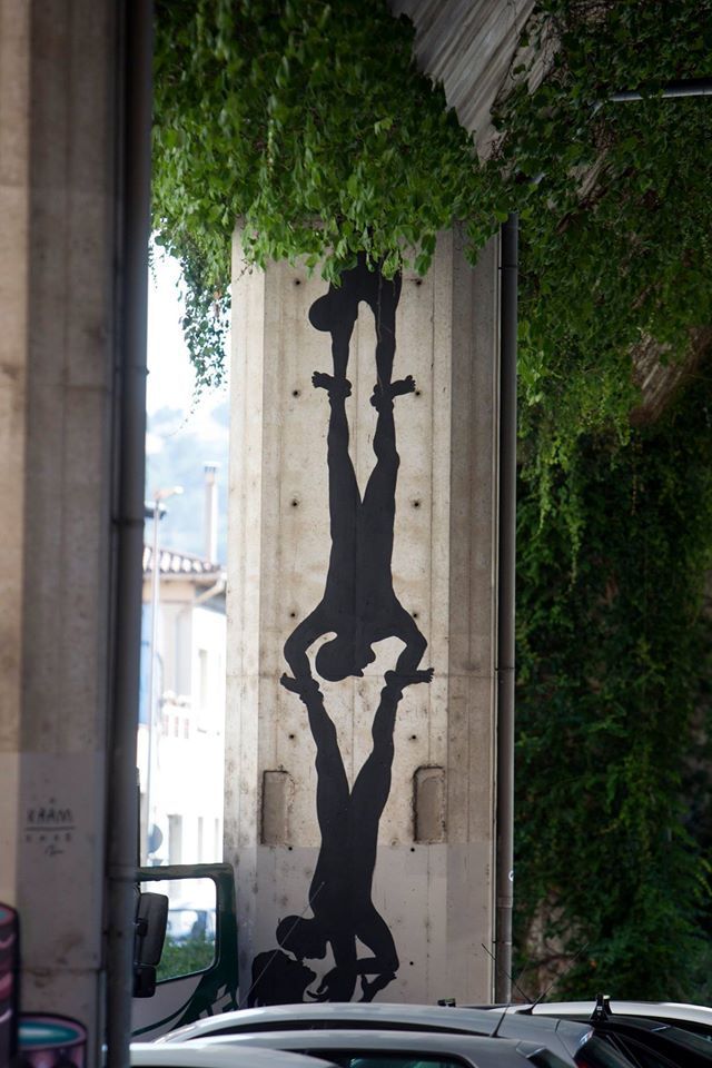 Mural of two black silhouette figures hanging upside down from a ledge, reaching down to grip the raised arms of two more figures below them, framed by hanging green vines and parked cars.