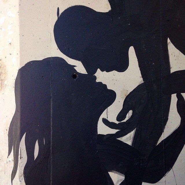 Close-up detail of a black silhouette mural showing two profile heads leaning in close together, one hand reaching up between them, painted on a wooden panel.