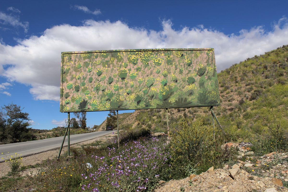 Roadside billboard entirely repainted as a dense camouflage pattern of green, tan, and yellow, blending it into the surrounding scrubland hillside, wildflowers growing at its base beside the road.
