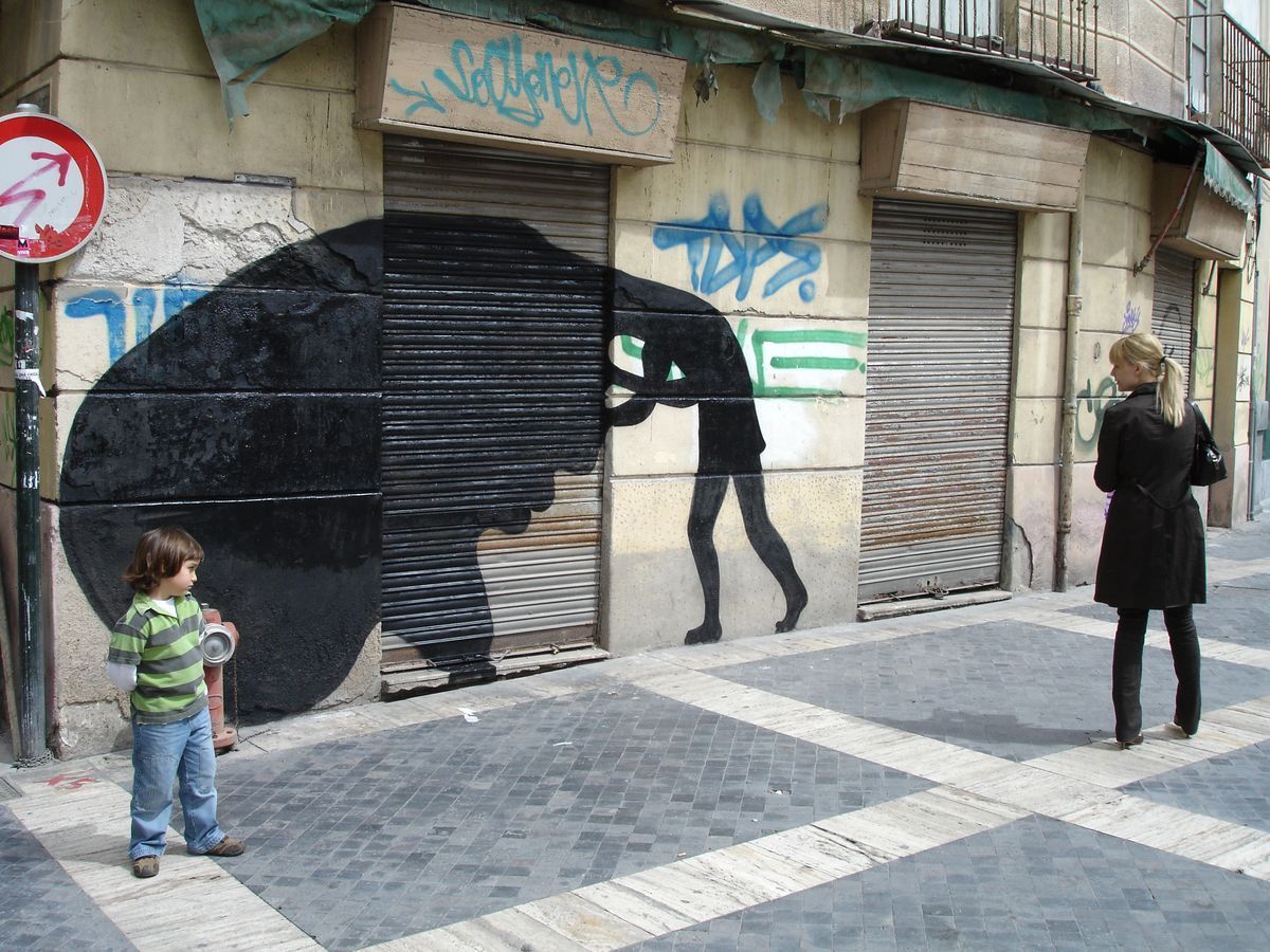 Black silhouette of a large head with an open mouth being pulled backward by a leg, facing a bird-like winged figure with dangling human legs, spray-painted graffiti above, an intercom button on the wall.