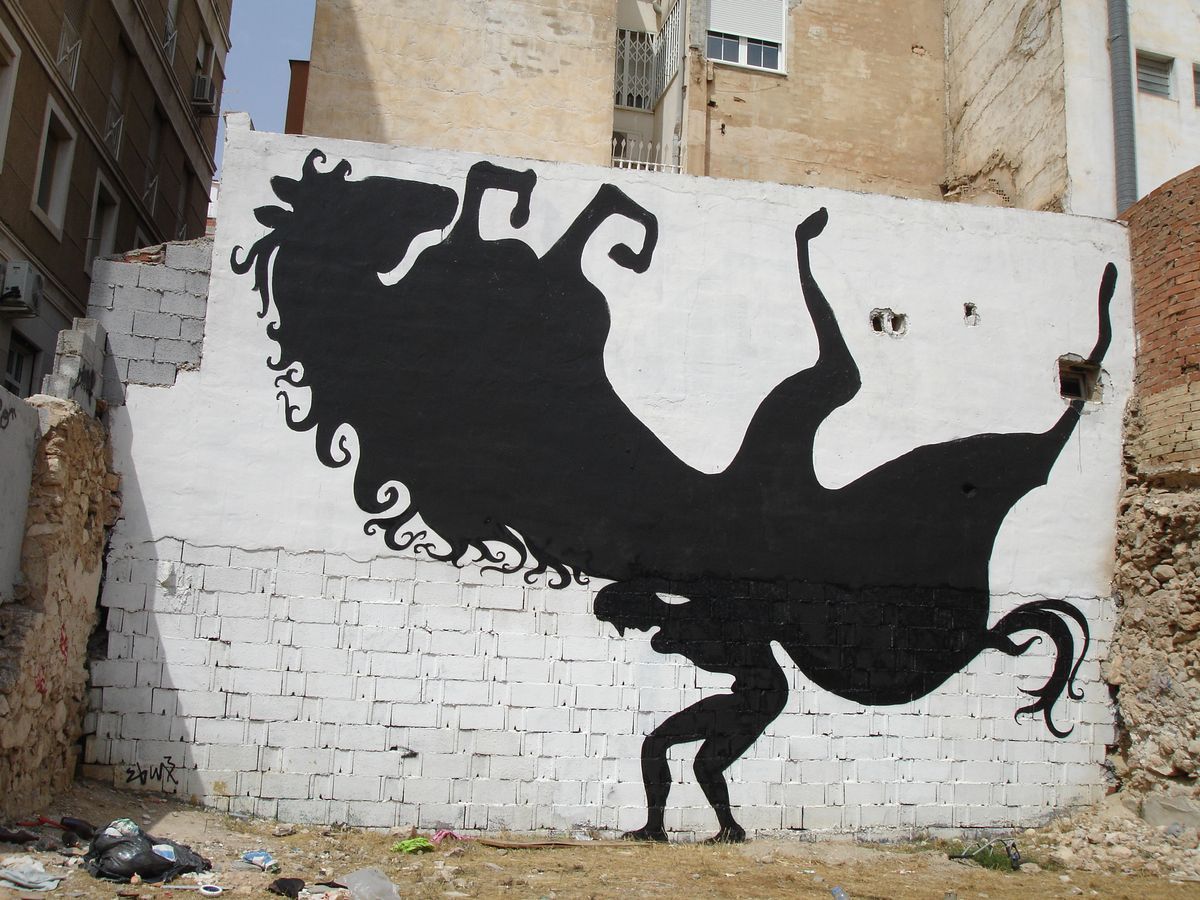 Black silhouette of a tall figure balanced on two long stilts held by two crouching figures pulling ropes at the base, painted on a ruined brick and plastered building corner.