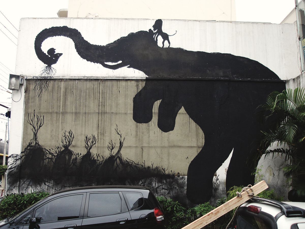 Large black silhouette of an elephant raising its trunk toward a small monkey, a tiny figure standing on its back, a row of leafless tree silhouettes below, painted on a building wall above parked cars.