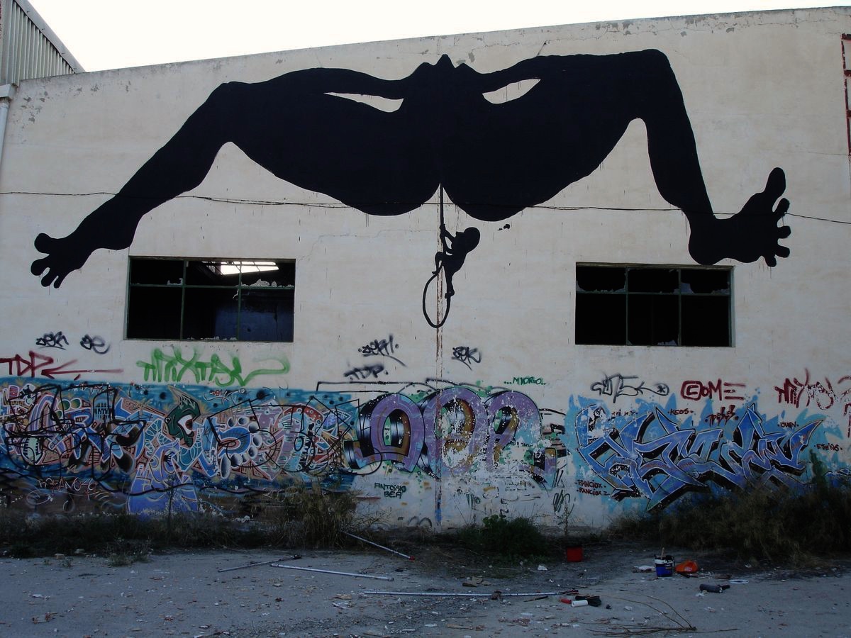 Giant black silhouette of an inverted nude figure sprawled horizontally across a warehouse facade, arms and legs spread wide, a tiny figure climbing down a rope from the crotch, graffiti covering the base of the wall.