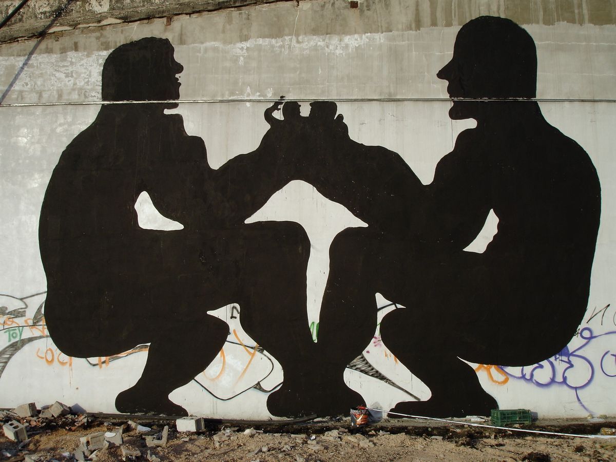 Two black silhouette figures squatting face to face, gripping a small trophy-like object between them with a white arrow shape pointing down beneath it, painted on a concrete wall covered in graffiti.