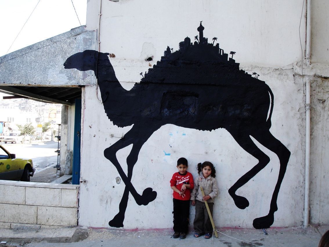 Black silhouette of a walking camel with a miniature hilltop city and minaret on its back, painted on a whitewashed wall, two children standing beneath it, a car parked to the side.